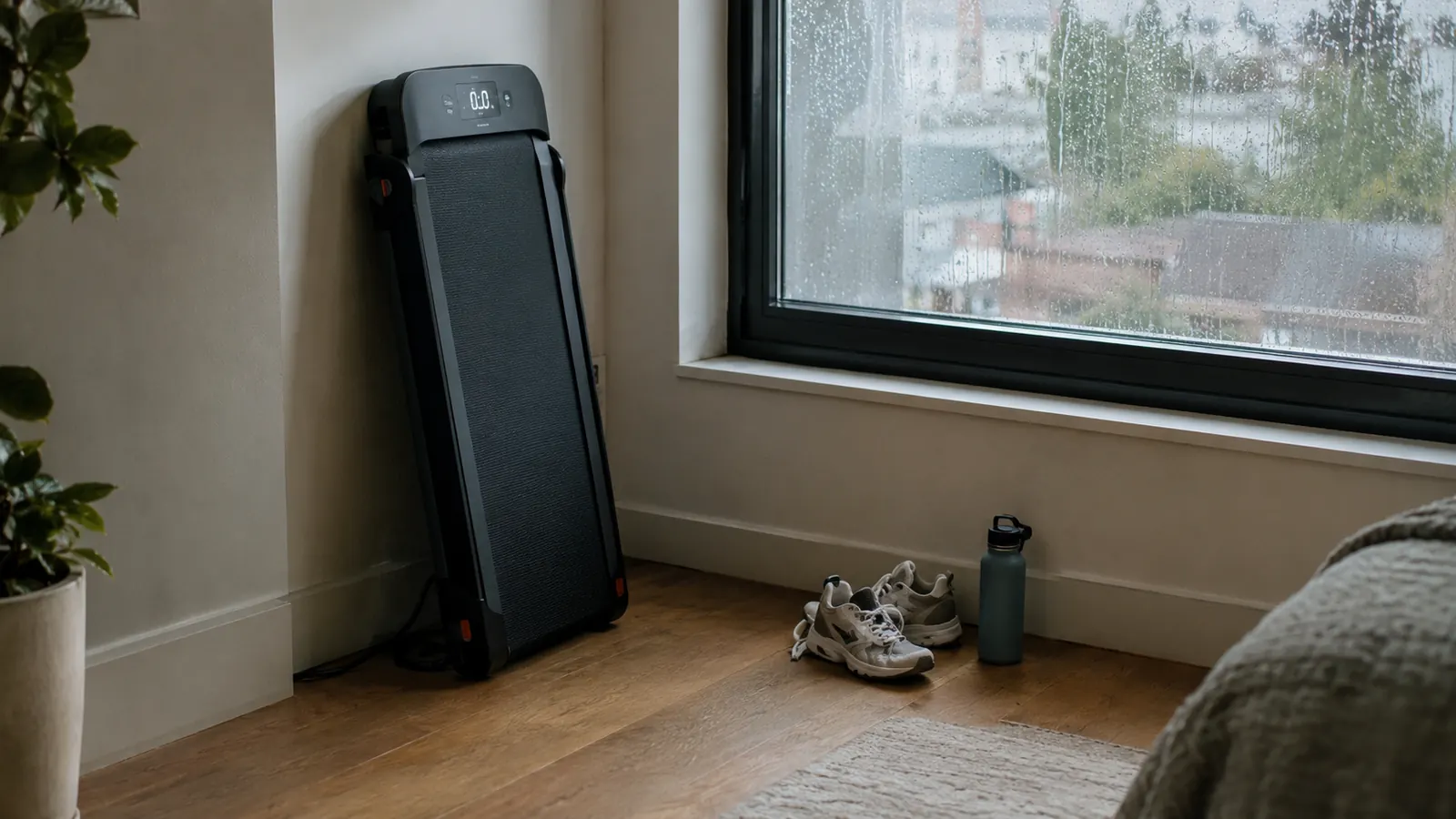 A compact walking pad in a spare room corner with rain on the window behind it
